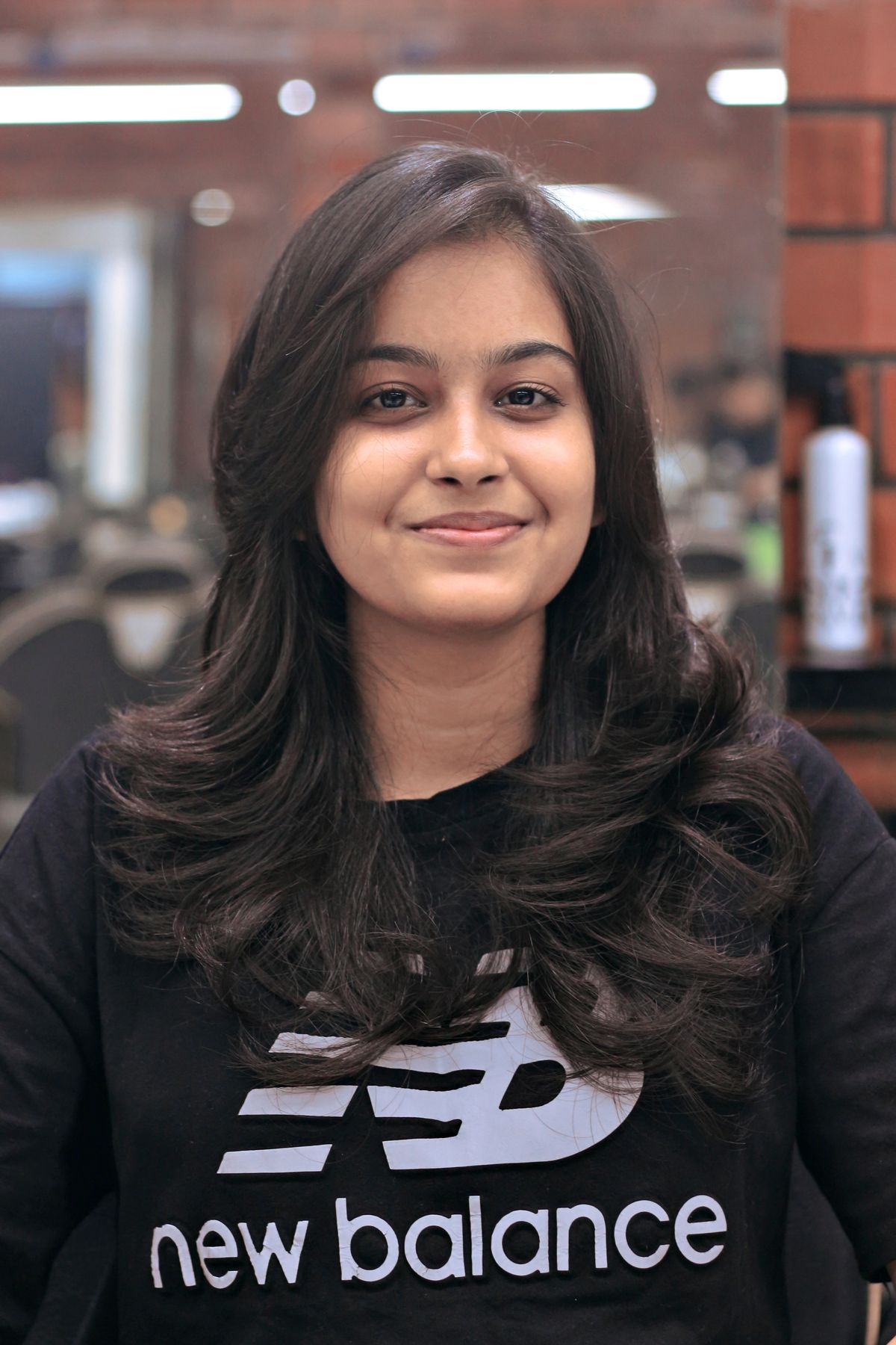 Young woman with brunette hair smiling in a salon wearing a fashionable sweatshirt.