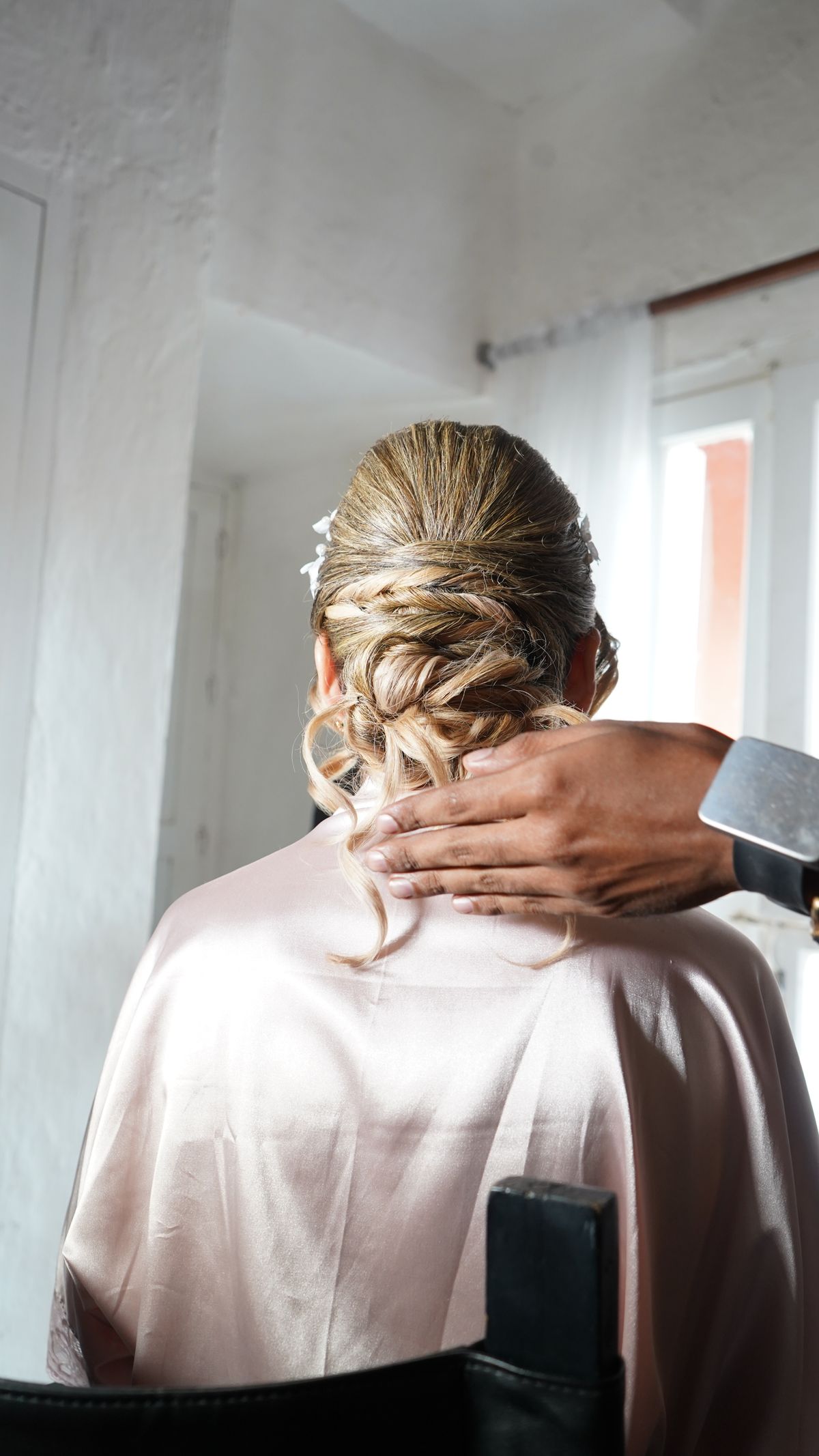 A stylist adjusts a bridal updo hairstyle in a well-lit room.